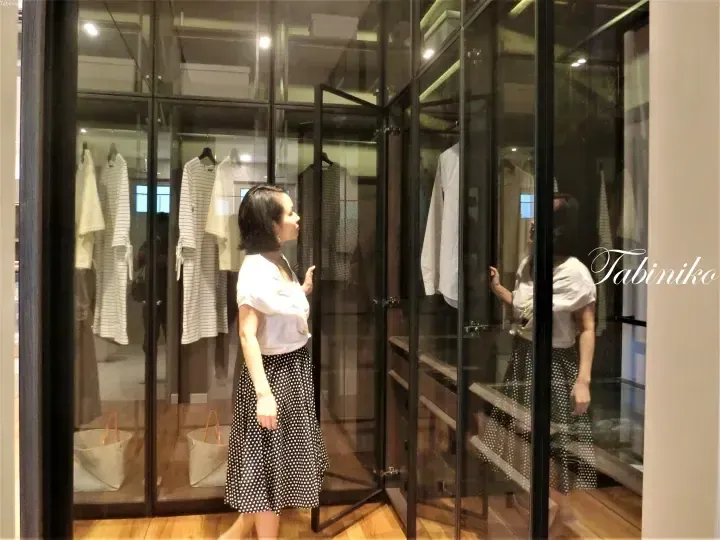Woman in polka dot skirt browsing clothes inside a modern glass walk-in closet with black frames.