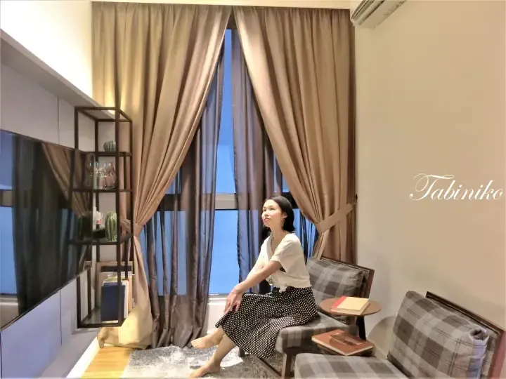 Woman relaxing in a modern apartment living room with large curtains, a TV, and a bookshelf.