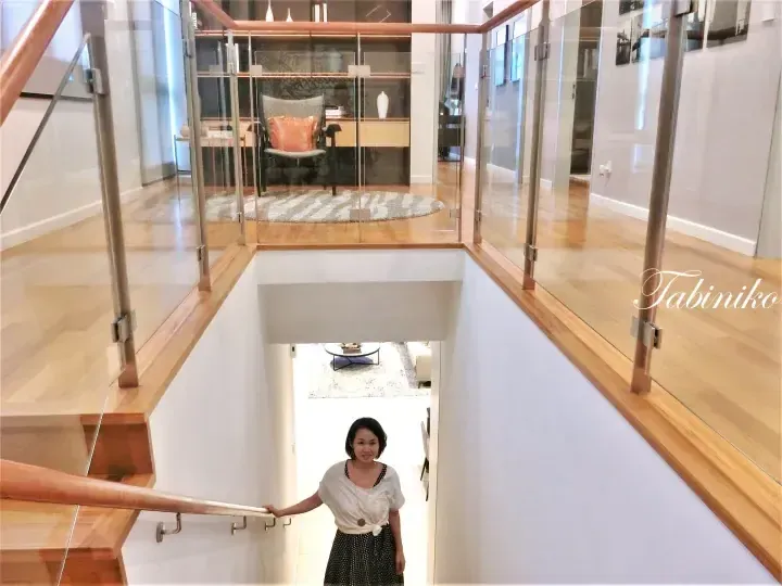 Woman standing on a modern wooden staircase with glass railings overlooking a bright open-plan living area.