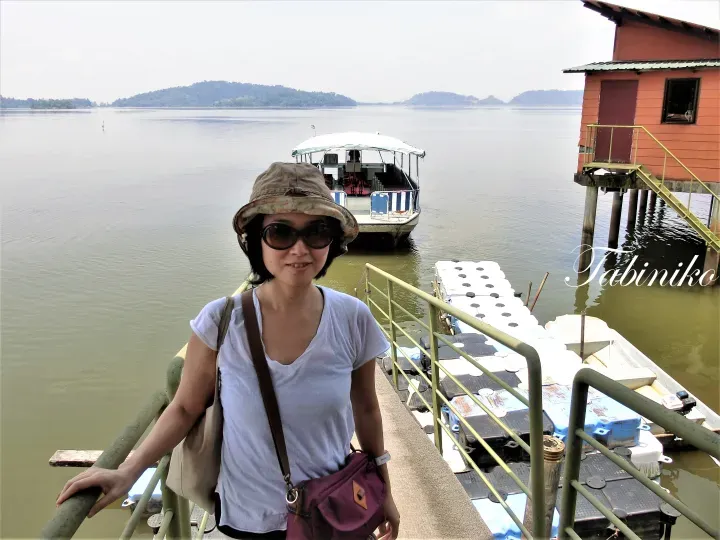 Woman in hat and sunglasses stands on a jetty at a lake resort with a tour boat and stilt house in the background.