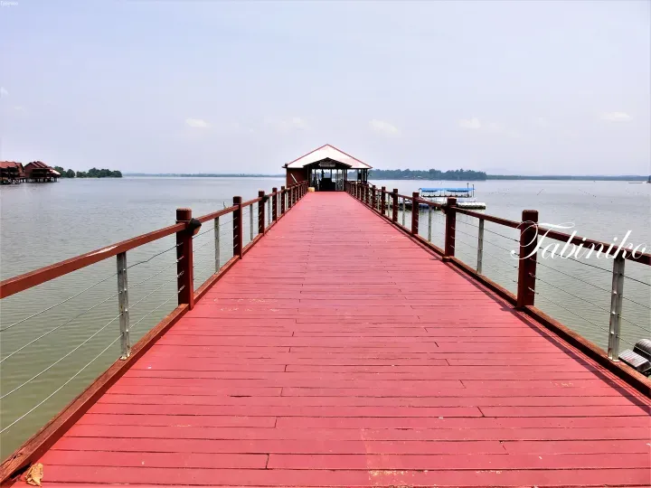 Long red wooden pier extending over calm water towards a ticketing counter at a resort.