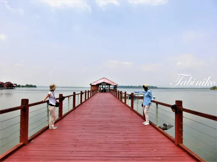 Two tourists on a red wooden pier overlooking a lake with a ticket counter building at the end.