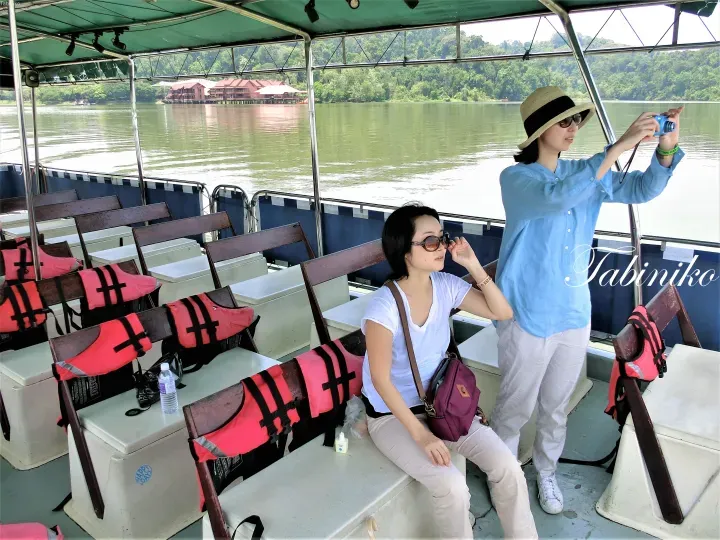 Two women on a boat tour, one taking a photo of the resort buildings along the riverbank.
