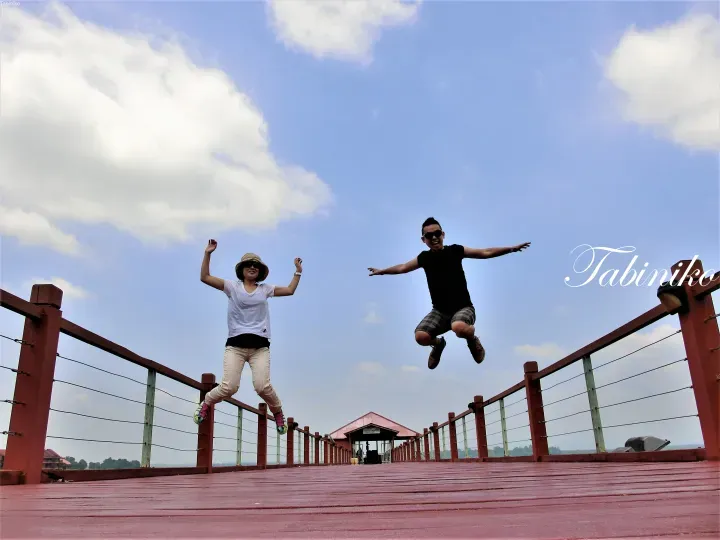 Couple jumping joyfully on a wooden pier at Tabiniko, with a blue sky and clouds in the background.