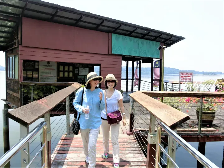 Two women walking on a wooden pier at Bukit Merah Orang Utan Island Foundation with a Departure sign and Malaysian flag.