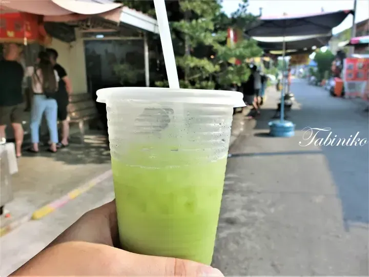 A refreshing green drink in a plastic cup held outdoors on a sunny street with blurred pedestrians and shops in the background.
