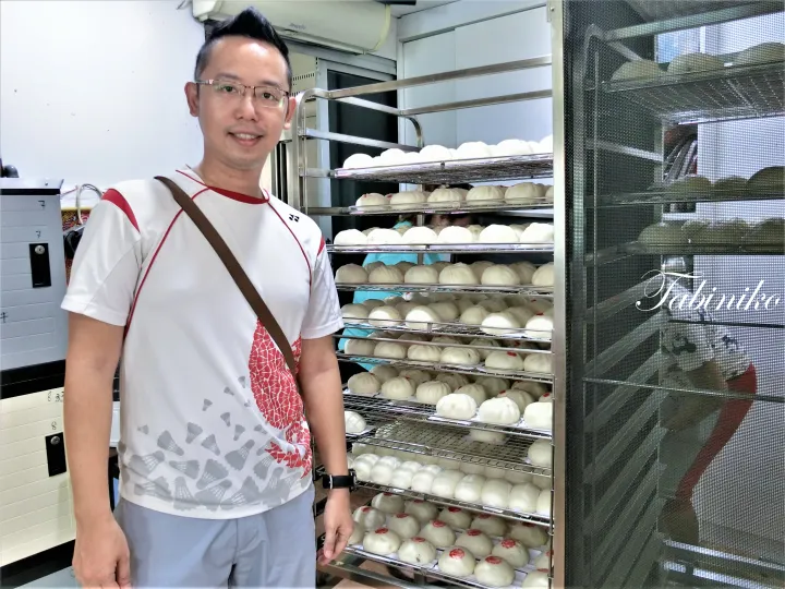 パン屋で、新鮮なパンが並んだ金属ラックの前に立つ男性。