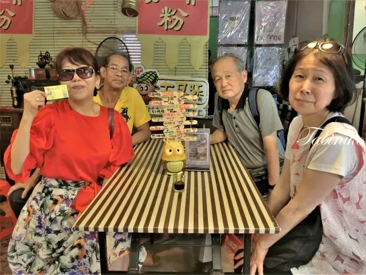 Four people pose at a striped table in a retro cafe, with a menu board displaying Kio Jia Coffee prices in the background.