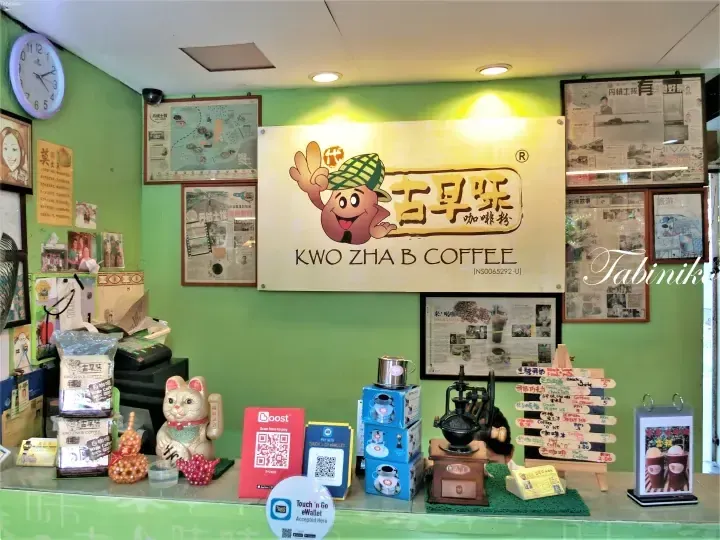 Kwo Zha B Coffee shop counter featuring a mascot sign, QR payment codes, and a vintage coffee grinder.