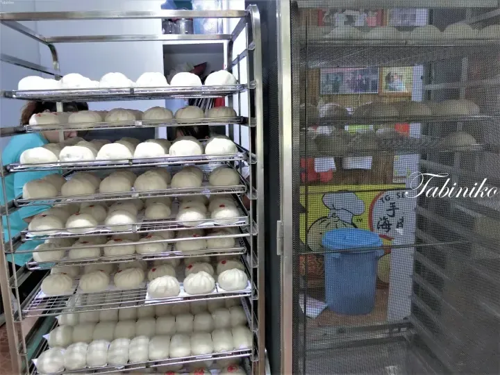 Rows of freshly steamed buns on metal racks inside a commercial kitchen, with a mesh door and blue bin visible.
