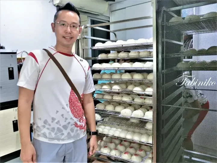 Man in white shirt standing beside metal racks filled with freshly made buns at a bakery.