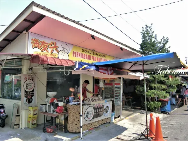 Hai Yew Heng shopfront in Kuala Langat displaying buns and coffee, with a worker behind the counter under a Malaysian flag.