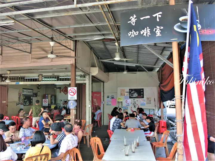マレーシア国旗が掲げられた屋外のカフェ「Tang Yi Xia Coffee Shop」で、赤いプラスチック椅子に座って食事をする客たち。