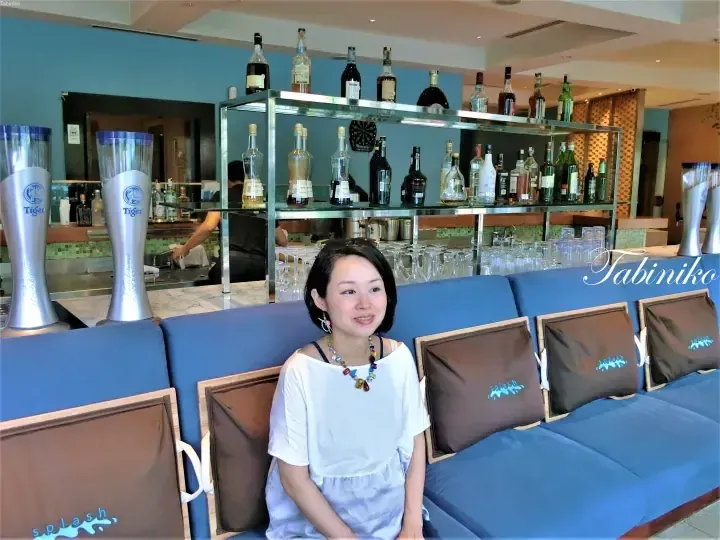 A woman sits on a blue sofa with Splash pillows at a bar counter featuring Tiger beer dispensers and liquor bottles.