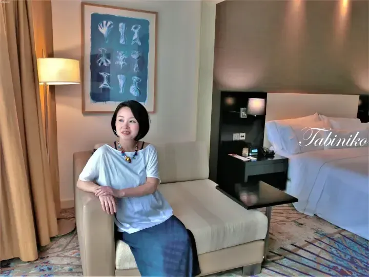 A woman sits on a beige sofa in a modern hotel room with a bed, artwork, and a lamp.