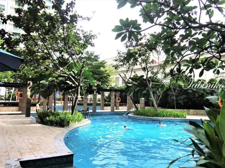 Winding swimming pool with blue tiles surrounded by lush tropical trees and a pergola at a resort.