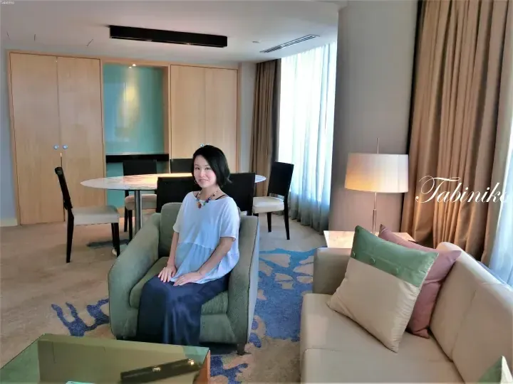 Woman sitting in a green armchair in a modern hotel suite living room with dining table and sofa.