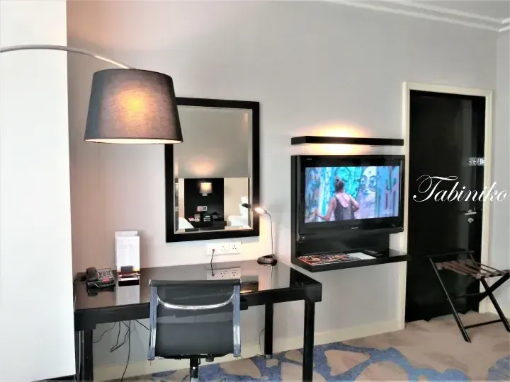 Modern hotel room interior featuring a black desk with a mirror, a flat-screen TV, and a luggage rack.