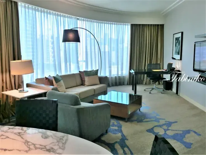 Modern hotel suite living room with sofa, armchair, glass coffee table, and large window overlooking city skyline.