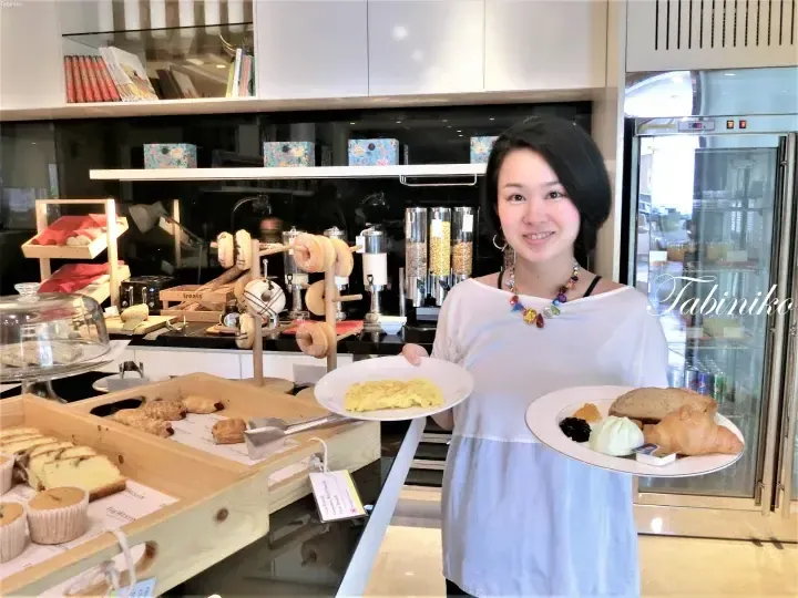 A woman holds plates of breakfast food at a buffet station featuring donuts, pastries, and an omelet at The Westin hotel.