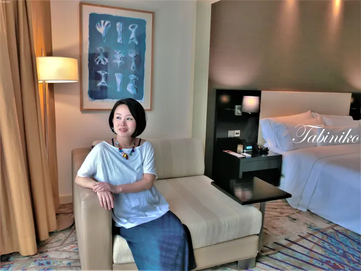 モダンなホテルの客室でソファに座る女性。ベッドやアート作品、ランプが見える。
