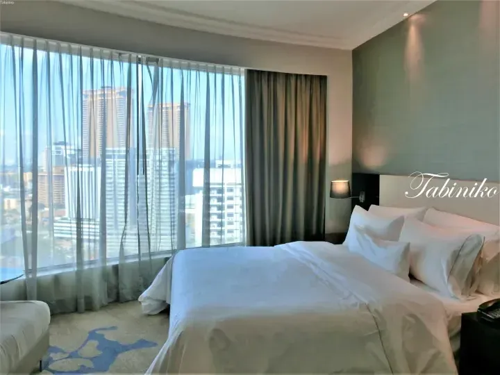 Spacious hotel room with a large bed and floor-to-ceiling windows offering a city skyline view.