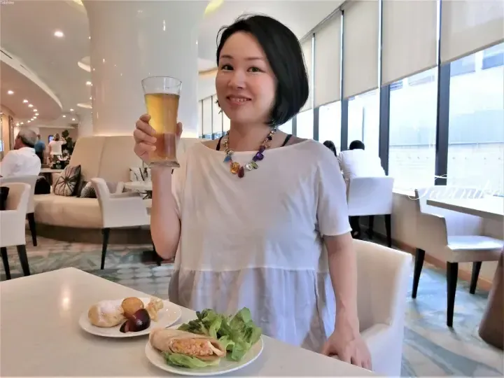 Woman holding a glass of beer in a bright hotel lounge with plates of food on the table.