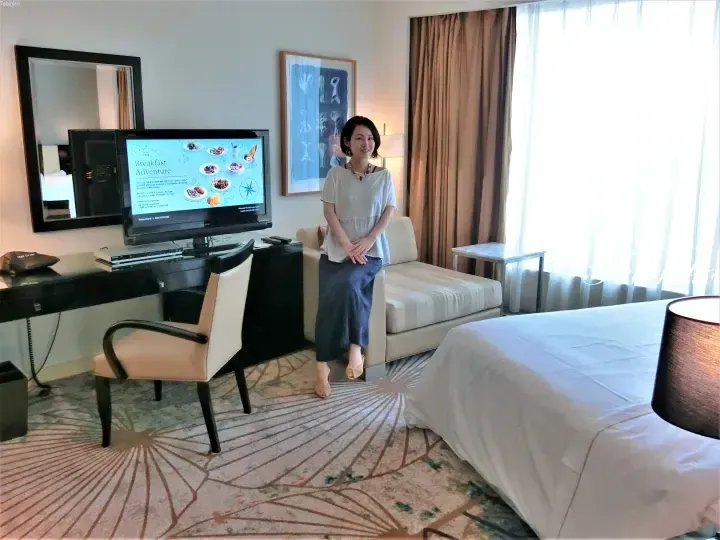 Woman standing in a hotel room near a sofa and TV displaying a breakfast menu.