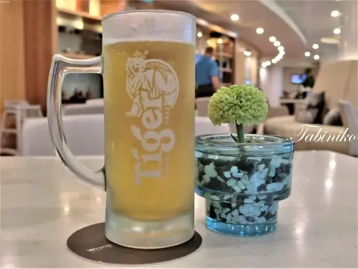 A frosty Tiger beer mug sits on a Westin coaster next to a small glass vase with a green flower.