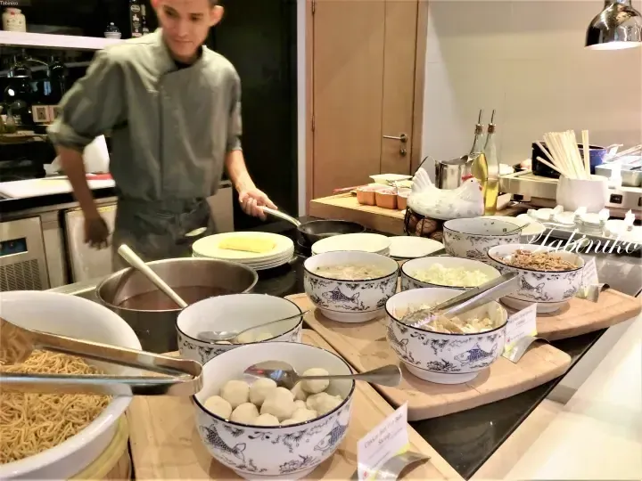 Chef preparing noodles at a buffet station with bowls of ingredients like fish balls and vegetables.