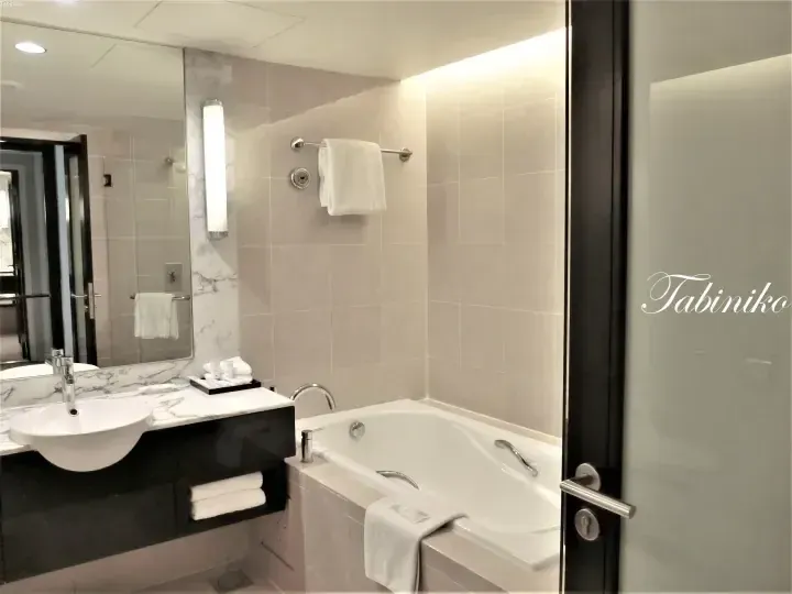 Modern hotel bathroom with marble vanity, sink, and bathtub featuring white towels and neutral tiled walls.