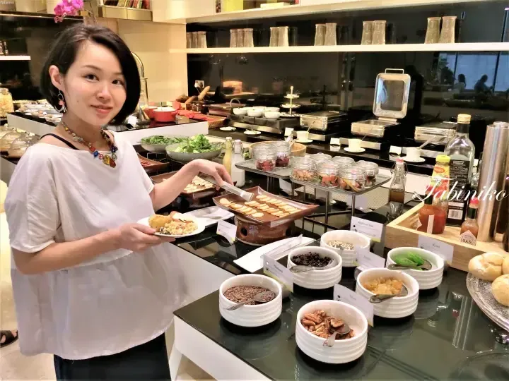 A woman selects food at a buffet station featuring bowls of nuts and seeds in a modern dining setting.