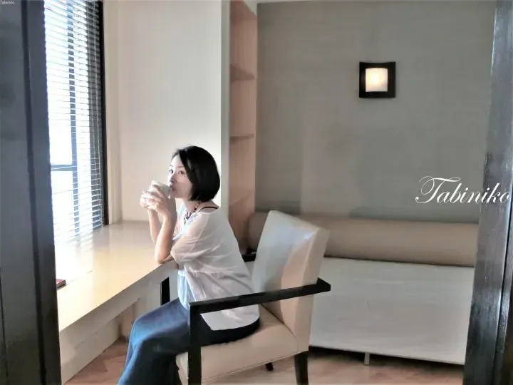 Woman in white shirt sipping coffee by window in modern hotel suite with sofa and wooden shelves