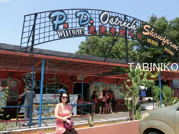 A woman stands in front of the P.D. Ostrich Showfarm entrance archway featuring Chinese characters and a red brick building.