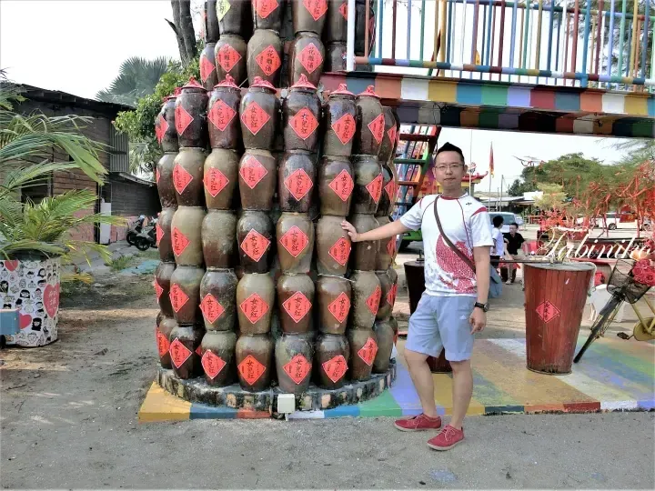Man posing beside a large stack of traditional wine jars at a Malaysian cultural site.