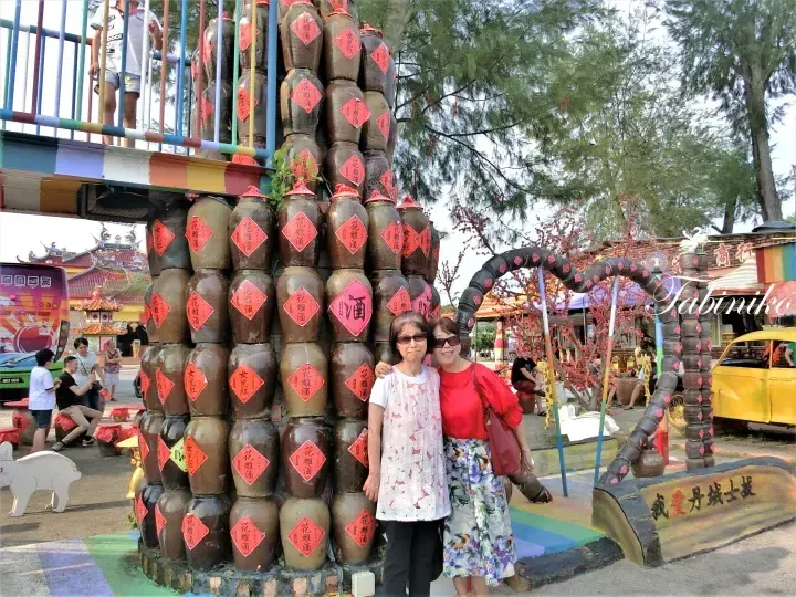 Two women pose in front of a large display of stacked ceramic jars labeled with Chinese characters at a Malaysian cultural site.