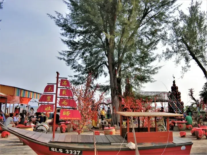 Red boat with Chinese banners at a beachside spot in Malaysia, surrounded by trees and visitors.