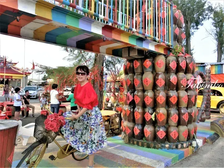 A woman in a red top poses with a bicycle near a colorful display of stacked clay jars at a Malaysian cultural attraction.