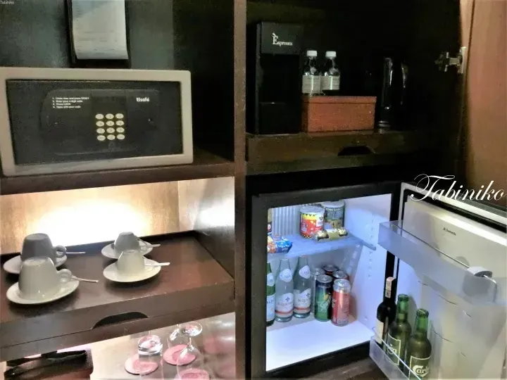 Hotel minibar with Dometic fridge stocked with Acqua Panna, Sprite, and Carlsberg, plus an Elsafe safe and espresso machine.