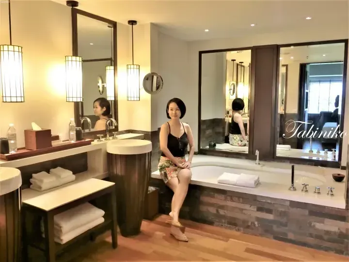 Woman sitting on the edge of a large bathtub in a modern luxury bathroom with wooden floors and elegant lighting.