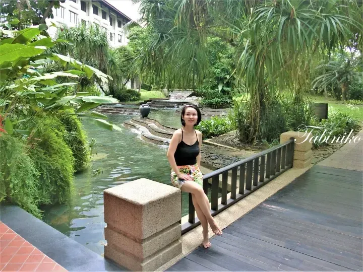 Woman sitting on a wooden bridge railing overlooking a resort water feature and lush tropical garden.