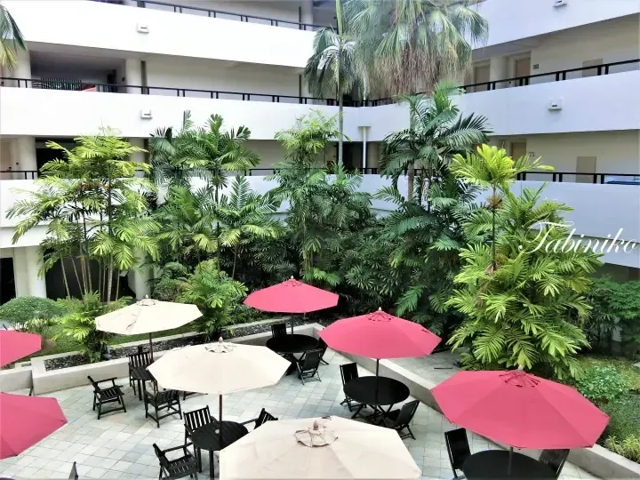 Outdoor seating area with red and beige umbrellas surrounded by lush tropical plants in a hotel courtyard.