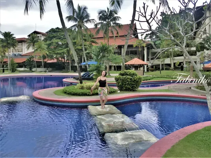 Woman standing on stepping stones in a resort pool with palm trees and a building with a red roof in the background.