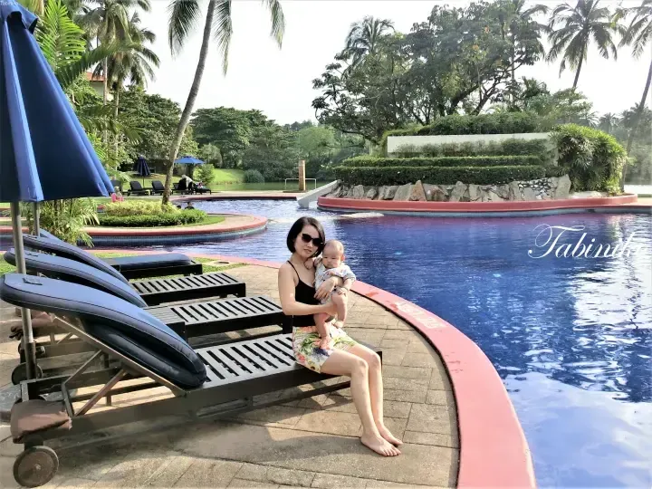 Woman holding a baby on a sun lounger beside a resort swimming pool surrounded by lush tropical greenery.