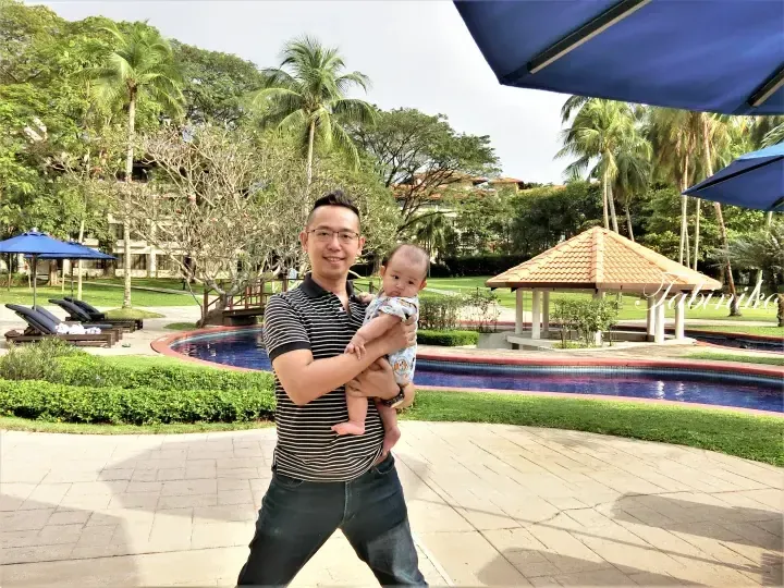 Man holding baby at resort poolside with gazebo, palm trees, and blue umbrellas in Malaysia.