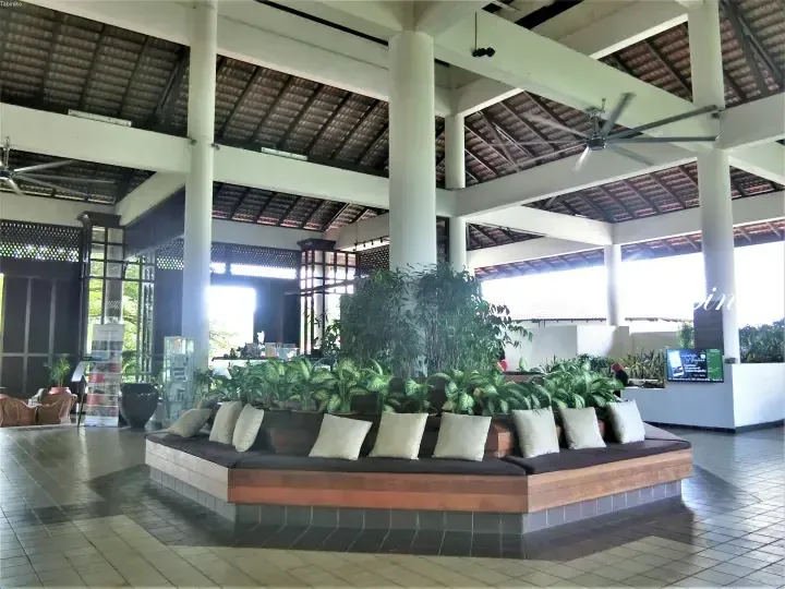 Spacious open-air lobby with high tiled ceilings, white pillars, and a large wooden daybed surrounded by lush green plants.