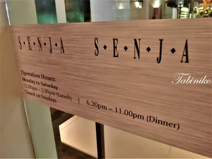 Wooden sign displaying operation hours for S·E·N·J·A restaurant, showing lunch and dinner times Monday to Saturday.