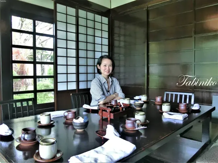 Woman seated at a long wooden table with tea sets in a traditional room with shoji screens and garden view