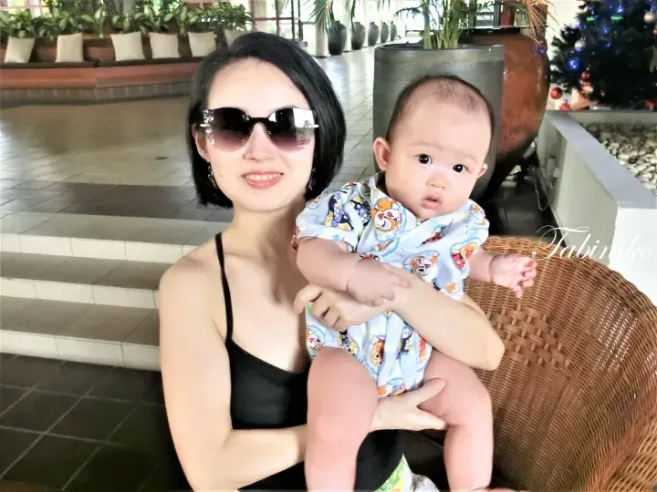 A woman wearing sunglasses holds a baby in a patterned shirt while sitting on a wicker chair in a resort lobby.