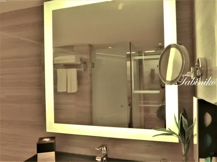 Modern hotel bathroom with backlit mirror, glass shower enclosure, and neatly arranged white towels.